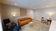This single-floor space showcases a thoughtfully designed living area that blends contemporary and mid-century modern aesthetics. The room is anchored by a rich cognac leather sofa paired with a striking sunburst-base glass coffee table, creating a sophisticated yet approachable seating arrangement. Complementing the sofa are accent chairs in deep navy and warm wood tones, adding visual depth to the space.

The walls are painted in a soft, neutral gray that provides a calm backdrop for three decorative wooden wall panels featuring intricate geometric and lattice patterns, serving as a standout artistic focal point. Light hardwood flooring runs throughout, enhancing the brightness of the room, while a multicolored abstract area rug in blue and gold tones grounds the seating area.

Recessed ceiling lighting provides clean, even illumination, supplemented by a contemporary globe floor lamp that adds a warm, ambient glow. White baseboards and clean wall lines contribute to a polished, well-finished interior. A white entry door is visible in the background, reinforcing the space's bright and airy character. Overall, the room exudes a modern, curated ambiance with a welcoming feel.