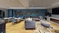 This thoughtfully designed clubhouse offers a welcoming and upscale communal environment across a single, open floor plan. Light hardwood flooring flows throughout the space, lending warmth and continuity to every zone.

The main lounge area is anchored by plush, tufted gray sofas and bold patterned accent chairs arranged around a central coffee table, inviting relaxed conversation and gathering. A striking large-scale sculptural wall installation adorns a deep teal feature wall, serving as a dramatic focal point alongside a fireplace. Geometric coffered ceiling details add architectural interest overhead, while recessed lighting ensures a bright, welcoming ambiance.

A dedicated seating nook near expansive floor-to-ceiling windows features a pair of rich blue velvet wingback chairs surrounding a gold-and-white pedestal table, offering an intimate retreat filled with natural light and outdoor views. Nearby, an elegant dining area accommodates a generous wood table surrounded by textured upholstered chairs, framed by dramatic patterned drapery cascading from the full-height glazing.

A central island counter area provides additional functionality, while lush indoor plants and curated decorative accessories contribute to the refined, contemporary aesthetic. The abundance of natural light streaming through the wraparound windows enhances the open, airy character of this versatile and beautifully appointed clubhouse.