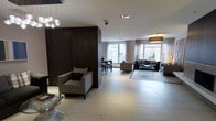 This stylish single-floor space is defined by its open-plan layout and refined contemporary design. The interior is anchored by large, light-filled windows dressed with sheer curtains, allowing natural light to flood the space and create a bright, airy atmosphere. The flooring throughout is finished in large-format light-toned tiles, lending a clean and cohesive look to the entire area.

A bold dark wood partition wall serves as a striking architectural feature, visually dividing the space into distinct functional zones while maintaining a sense of openness. The living area is thoughtfully arranged with plush upholstered seating, including armchairs and a sofa ensemble, complemented by a sleek glass-and-metal coffee table resting on a textured area rug. A warm pendant lamp adds an inviting glow to this lounge zone.

Adjacent to the living area, a casual dining nook features bar-height seating at a narrow table, ideal for informal gatherings. The space is further enhanced by a modern cluster pendant chandelier with globe lights, adding a decorative focal point near the dining area.

Throughout, the design palette combines warm wood tones, neutral upholstery, and metallic accents, creating an atmosphere that is both sophisticated and welcoming — well-suited for both relaxed living and stylish entertaining.