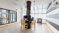 This single-floor space serves as a well-appointed lobby and leasing office, thoughtfully designed with a modern aesthetic that balances bold visual elements with functional areas. The entrance greets visitors with a striking accent wall featuring broad, alternating black and white horizontal stripes, illuminated by wall-mounted sconce lighting and complemented by warm stone tile flooring that carries throughout the space.

At the heart of the lobby stands a distinctive circular workstation wrapped around a glossy black cylindrical column. Equipped with desktop computers and bar-height wooden seating, this central hub is ideal for resident or guest use. Overhead, chain-hung pendant lights with Edison-style bulbs add an industrial-chic touch that contrasts elegantly with the clean white ceilings.

A standout feature is the sunroom-style seating alcove, enclosed by floor-to-ceiling arched glass panels that flood the area with diffused natural light. Two plush, dark wingback chairs with striped accent pillows flank a wire-frame side table, creating an inviting lounge atmosphere.

Adjacent areas include a practical service corridor with dark cabinetry, a printer station, and interior glass partitions that offer views into a separate workspace or conference area. Large sliding glass doors provide a visual connection to an outdoor courtyard with additional seating, reinforcing the open, airy character of the overall space.