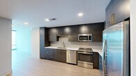 Located in the heart of downtown Raleigh, this high-rise unit on the 14th floor offers a thoughtfully designed one-bedroom, one-bathroom layout with a clean, modern aesthetic. Light wood-tone flooring flows seamlessly throughout the space, complementing the neutral palette of white and soft gray walls.

The open kitchen is a standout feature, outfitted with two-tone cabinetry — rich dark wood uppers paired with deep teal-toned lowers — white quartz countertops, a subway tile backsplash, and a full suite of stainless steel appliances including a gas range, built-in microwave, dishwasher, and French door refrigerator with ice dispenser.

The living area benefits from expansive floor-to-ceiling windows that frame striking panoramic views of the Raleigh skyline, flooding the space with abundant natural light. The bedroom continues this design language with its own floor-to-ceiling window wall offering city views, a dedicated closet, and access to the full bathroom.

The bathroom features large-format tile flooring, a soaking tub with shower, a wide single-sink vanity with contemporary cabinetry, and recessed lighting. A dedicated closet completes the unit's functional layout. The overall ambiance is sleek, bright, and urban — ideal for those seeking a modern downtown lifestyle.