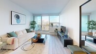 This well-designed one-bedroom, one-bathroom unit offers a smartly efficient layout across a single floor, maximizing every square foot with modern finishes and thoughtful details throughout. The open-concept living and kitchen area serves as the heart of the unit, where light hardwood-style flooring and crisp white walls create a clean, contemporary aesthetic. The living room is anchored by expansive floor-to-ceiling windows that flood the space with natural light and frame sweeping city skyline views, with access to a balcony beyond. The kitchen is outfitted with white shaker cabinetry, a stainless steel appliance suite including a full-size refrigerator, range, and over-the-range microwave, and a peninsula with bar seating for two — ideal for casual dining and entertaining. The bedroom is a serene retreat featuring wide windows with city views, a built-in sliding-door closet with shelving, and continuity of the light hardwood flooring. A hallway connects the bedroom to the bathroom, which features a light wood vanity with an undermount sink, white quartz countertop, a large mirror, and clean tile flooring. The unit's consistent white palette and modern fixtures create a cohesive, airy atmosphere throughout.