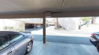 This multi-unit residential complex is organized around a central courtyard layout, offering a practical and functional design suited for community living. The exterior presents a classic stucco finish in neutral tones, characteristic of Southern California-style construction, with buildings spanning two stories connected by exterior stairways.

The ground level features a generously sized covered carport area with designated parking spaces supported by sturdy concrete columns. The carport provides shade and protection from the elements, accommodating multiple vehicles with ease. Beyond the parking area, a wide concrete drive lane runs through the property, providing clear and accessible circulation throughout the complex.

The courtyard space is highlighted by mature trees that offer natural shade and a touch of greenery, complemented by neatly maintained shrubs and landscaping along building edges. Exterior stairways with metal railings provide access to the upper-level units, while walkways and balconies line the building facades.

The overall ambiance of the property is practical and well-maintained, with an open-air design that allows for ample natural light and airflow throughout the common areas. The layout fosters an accessible and connected environment, making it a straightforward and functional residential community.