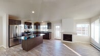 This single-story home is designed with an open, flowing layout that connects the kitchen and living areas seamlessly. The kitchen is a standout feature, showcasing rich dark-stained cabinetry that extends to the vaulted ceiling, contrasting elegantly with dark granite countertops. A central island with matching granite provides additional workspace and serves as a natural gathering point. Stainless steel appliances, including a refrigerator, range, and microwave, complete the kitchen's modern aesthetic. Multiple windows allow generous natural light to fill the space, enhancing its airy feel.

The living area adjoins the kitchen and is anchored by a built-in fireplace set into a clean white surround, adding warmth and a focal point to the room. Vaulted ceilings elevate the sense of space throughout the main living areas. Light wood-look flooring runs consistently across the home, lending a cohesive, contemporary feel.

The primary bedroom features soaring ceilings, abundant natural light, a ceiling fan, and direct access to a walk-in closet and an en suite bathroom with dark cabinetry and a vanity sink. Additional bathrooms include tub-shower combinations with chrome fixtures and light wood-look flooring, maintaining a clean, neutral palette throughout.