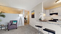 This single-floor residence offers a thoughtfully designed layout that balances comfort and functionality. The open-concept kitchen is a standout feature, outfitted with gray shaker-style cabinetry, granite countertops, and black appliances including a full-size refrigerator and range. A peninsula breakfast bar with bar seating extends into the living area, creating a natural transition between cooking and entertaining spaces. A large window floods the kitchen with natural light, complementing the wood-look flooring underfoot.

The living room is generously sized with plush wall-to-wall carpeting and features a distinctive decorative upper partition that adds architectural character to the space. A wall-mounted HVAC unit provides climate control, and the textured ceiling adds a classic touch throughout.

The bedroom is comfortably proportioned with soft carpeting, a high clerestory-style window fitted with vertical blinds, and ample room for a full furniture arrangement including a desk area, making it suitable as a dual-purpose sleeping and workspace.

The bathroom is well-appointed with a gray vanity, granite countertop, an undermount sink, and a large mirror. Clean white walls and teal accents create a bright, refreshing atmosphere. The overall ambiance of the space is warm, approachable, and move-in ready.