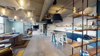 This single-floor communal space blends industrial and contemporary design into a dynamic, multi-functional environment. Polished concrete floors run throughout, complementing exposed ductwork and piping overhead that lend the space an open, loft-like character. Bold teal and navy blue tones anchor the design, featured prominently in a striking chevron-patterned acoustic wall panel that adds both visual interest and sound absorption to the dining and lounge areas.

The layout is thoughtfully segmented into distinct zones. A café-style dining area offers an abundance of mixed seating, including light wood chairs at communal tables and counter-height bar stools along a sleek navy kitchenette fitted with open shelving and a sink. Large windows flood this area with natural light and provide views to the outdoors. An adjacent lounge zone features a generous sectional sofa, patterned area rugs, and statement pendant lighting, creating a relaxed, residential atmosphere.

A long built-in wooden bench with cushions lines one wall, offering casual seating alongside small café tables with colorful chairs. Further into the space, a more private lounge area includes a large-screen display and additional soft seating. Deep teal built-in shelving units and locker-style storage elements add both function and bold color. Throughout, sculptural pendant lights and geometric ceiling fixtures contribute to an energetic yet welcoming ambiance.