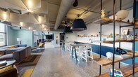 This single-floor communal space blends industrial and contemporary design into a dynamic, multi-functional environment. Polished concrete floors run throughout, complementing exposed ductwork and piping overhead that lend the space an open, loft-like character. Bold teal and navy blue tones anchor the design, featured prominently in a striking chevron-patterned acoustic wall panel that adds both visual interest and sound absorption to the dining and lounge areas.

The layout is thoughtfully segmented into distinct zones. A café-style dining area offers an abundance of mixed seating, including light wood chairs at communal tables and counter-height bar stools along a sleek navy kitchenette fitted with open shelving and a sink. Large windows flood this area with natural light and provide views to the outdoors. An adjacent lounge zone features a generous sectional sofa, patterned area rugs, and statement pendant lighting, creating a relaxed, residential atmosphere.

A long built-in wooden bench with cushions lines one wall, offering casual seating alongside small café tables with colorful chairs. Further into the space, a more private lounge area includes a large-screen display and additional soft seating. Deep teal built-in shelving units and locker-style storage elements add both function and bold color. Throughout, sculptural pendant lights and geometric ceiling fixtures contribute to an energetic yet welcoming ambiance.