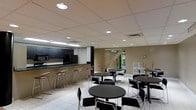 This single-floor space functions as a well-appointed communal lounge and kitchenette area, designed to accommodate a variety of gathering and dining needs. The layout is open and spacious, allowing for easy movement between the seating area and the service counter. The dropped ceiling features a clean grid of recessed pot lights and flush fluorescent fixtures, providing consistent, even illumination throughout the space.

The seating area is furnished with multiple round black tables paired with sleek chrome-framed chairs, arranged to support both small group interactions and larger gatherings. Bar-height seating lines the counter along one wall, offering an additional informal dining option. The kitchenette runs along the back wall and is outfitted with dark cabinetry, a long dark countertop, and visible appliances, making it suitable for food preparation and service.

Light-toned ceramic tile flooring extends throughout, contributing to the bright and clean aesthetic of the space. Warm decorative branches in vases add subtle visual interest. A doorway at the rear provides a glimpse into an adjacent area, suggesting connectivity to further spaces within the building.