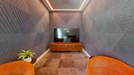 Located at 100 Hamilton St in Bound Brook, New Jersey, this contemporary game room is a thoughtfully designed amenity space that blends entertainment and relaxed socializing. The open single-level layout is finished with large-format tile flooring in a soft grey palette, creating a clean and modern foundation throughout.

The space is divided into distinct activity zones. A vibrant arcade section lines one wall, housing a collection of classic upright arcade cabinets — including titles like Pac-Man, Wheel of Fortune, and others — offering a nostalgic yet lively gaming experience. Nearby, a full-size foosball table anchors the central area, providing a focal point for competitive play.

A recessed media lounge nook adds a more intimate dimension to the room. Clad in richly textured, geometric-patterned wall panels with warm ambient cove lighting above, it features a wall-mounted flat-screen television, a sleek wooden media console, and plush orange accent chairs — creating a cozy retreat within the larger space.

Floor-to-ceiling windows along one wall flood the room with natural light and offer views to the exterior, while a high-top bistro table and seating provide a casual spot for conversation. Recessed lighting and continuous cove lighting along the ceiling perimeter maintain a warm, inviting ambiance throughout the entire space.