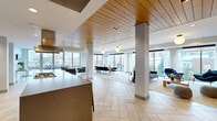 Located at 315 First Ave NE in Minneapolis, this sophisticated single-floor amenity space offers a thoughtfully designed open-plan layout that seamlessly integrates multiple functional zones. The interior is anchored by striking herringbone-patterned stone tile flooring and a warm wood-slatted ceiling that adds texture and visual depth throughout.

The fully equipped kitchen serves as a centerpiece, featuring a large island with a built-in gas cooktop, a stainless steel hood vent, dark cabinetry, open yellow shelving against a geometric patterned tile backsplash, and bar seating for casual gathering. Adjacent to the kitchen, a long dining table with modern black chairs is positioned to take full advantage of the floor-to-ceiling windows lining the perimeter.

Multiple lounge seating areas are arranged throughout the space, offering flexibility for both relaxed socializing and more structured gatherings. One lounge area is anchored by a sleek linear fireplace set into a textured stone wall, topped by a large-screen TV. Sheer drapery softens the abundant natural light streaming in from the expansive windows.

A dedicated seating nook provides direct access to an outdoor terrace through tall glass doors, offering views of the Minneapolis skyline. The overall ambiance is modern and refined, blending warm materials with contemporary design for an inviting, upscale communal experience.