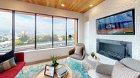 This thoughtfully designed single-floor space showcases a seamless open-concept layout that integrates the kitchen, dining, and living areas into one cohesive environment. The kitchen is a standout feature, anchored by a striking live-edge wood island with a light blue base and seating for multiple guests. White cabinetry and stainless steel appliances line the walls, while a bold, colorful mosaic backsplash adds artistic energy to the space. Pendant lights with gold interiors cast a warm glow overhead, complementing the large-format tile flooring throughout.

The living area is defined by a textured white fireplace wall with a mounted television, paired with contemporary furnishings and a vibrant teal-accented area rug. Large floor-to-ceiling windows flood the room with natural light and frame sweeping city and bridge views, while a warm wood-paneled ceiling adds architectural warmth and contrast. The dining area is positioned near French doors that open directly onto a furnished outdoor terrace, extending the living space and offering additional cityscape views. Overall, the space balances modern design with curated, artistic details for a refined yet inviting atmosphere.