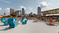 Located at 1254 Hennepin Ave N in Minneapolis, this rooftop terrace offers a thoughtfully designed outdoor amenity space with panoramic views of the downtown Minneapolis skyline. The terrace is divided into several distinct zones, each serving a specific purpose while contributing to a cohesive and vibrant outdoor environment.

A full-length bocce ball court sits at the heart of the terrace, bordered by brick edging and framed by lush artificial turf. Alongside the court, a sun-bathing area features a row of white lounge chairs, ideal for relaxing while taking in the surrounding cityscape. Scattered seating throughout the turf areas includes modern white lounge furniture with accent tables, providing informal gathering spots.

A covered outdoor kitchen and dining area is equipped with grills, bar-height tables, and colorful seating, making it well-suited for outdoor entertaining. Adjacent to this, a fire pit lounge with bold teal modern chairs creates a striking focal point against the concrete terrace surface.

Raised planter beds throughout the space are filled with flowering plants, ornamental shrubs, evergreen trees, and sculptural greenery, adding color and a garden atmosphere to the urban setting. Perimeter railings provide unobstructed sightlines to the Minneapolis skyline on all sides, making this rooftop terrace a standout outdoor amenity in the heart of the city.