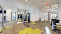 Located at 2818 Barrow Rd. in Charlotte, NC, this single-floor fitness center is designed to support a comprehensive workout experience in a bright, modern environment. The space features soaring ceilings that lend an open, airy atmosphere, complemented by an abundance of large windows that allow generous natural light to flow throughout and offer views of the surrounding neighborhood.

The fitness center is divided into well-organized workout zones. The cardio area is anchored by a row of treadmills and elliptical machines, each equipped with personal display screens and positioned along a dramatic dark accent wall with clerestory windows above and a wall-mounted flat-screen TV. Adjacent to this, a dedicated strength training area houses a cable machine, a multi-station weight system, a dumbbell rack with bench, and a stair climber, providing ample options for resistance and functional training.

A full-length wall mirror in the strength zone enhances the sense of space while allowing users to monitor their form. The flooring is a stylish combination of patterned carpet tiles accented with bold yellow sections, adding a dynamic visual energy to the space. Ceiling fans and recessed lighting ensure comfort throughout. Glass-panel entry doors contribute to the open, accessible feel of this thoughtfully appointed community fitness facility.