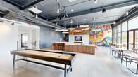 This expansive single-floor amenity facility offers a thoughtfully designed mix of social, recreational, and professional spaces. The centerpiece is the "G Lounge," a striking open-plan social area featuring polished concrete floors, an exposed dark ceiling with industrial ductwork, and dramatic pendant chandelier fixtures. A full-length bar counter with warm wood paneling and barstool seating anchors the space, while a shuffleboard table invites casual recreation. Bold, large-scale murals add vibrant artistic energy throughout.

An adjacent bright lounge area opens to the outdoors through expansive floor-to-ceiling windows, flooding the space with natural light and offering views of the surrounding landscape. This area features a kitchenette with a sink, mixed seating arrangements on a plush area rug, and decorative shelving with curated décor.

The well-equipped fitness center includes a full array of cardio machines, free weights, cable machines, and strength equipment, all set against floor-to-ceiling mirrors with a clean, bright aesthetic.

A private office area with carpeted flooring, a dropped ceiling, and warm wood furnishings provides a quiet, professional environment. Throughout the space, the design balances industrial and contemporary elements to create an inviting, multifunctional destination.