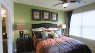 The bedroom is a standout feature of this single-story space, combining rich color and classic design elements to create a warm, inviting atmosphere. The walls are painted in a deep sage green, providing a bold yet grounding backdrop that pairs seamlessly with the dark espresso-toned furnishings throughout the room. A substantial upholstered headboard anchors the bed, which is dressed in layered earth-toned bedding featuring shades of rust, brown, and gray, accented by an assortment of patterned and textured throw pillows. Flanking the bed are matching dark wood nightstands, each topped with warm-toned table lamps that contribute to the room's cozy ambiance. A trio of ornate framed artwork is displayed symmetrically above the headboard, adding a decorative focal point to the space. Natural light enters through a window dressed with dark curtains and horizontal blinds, offering both light control and privacy. A ceiling fan with an integrated light fixture is centrally positioned overhead, ensuring comfort and functionality. The overall aesthetic is cohesive, blending traditional design sensibilities with rich, warm tones throughout.