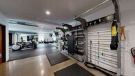 This fully equipped fitness center is thoughtfully designed to support a wide variety of workout routines. The space is divided into distinct training zones, each optimized for specific exercise disciplines. The cardio section features a row of elliptical trainers, treadmills, and stationary bikes, including spin bikes, all positioned near windows that allow natural light to fill the room and provide views of the surrounding exterior. Full-length wall mirrors amplify the sense of space and allow users to monitor their form during exercise. Overhead flat-screen televisions are mounted throughout, enhancing the workout experience. The strength and functional training area is anchored by a multi-use TRX and resistance training rig, stocked with resistance bands, suspension trainers, and a variety of attachments. A dedicated free weights section includes a well-organized dumbbell rack alongside flat and adjustable benches. A Rogue-branded flat bench and an Endurance rowing machine round out the strength offerings. Rubber flooring and exercise mats provide safety and comfort underfoot. Recessed lighting throughout ensures even illumination, while exposed ductwork adds an industrial design element. The facility is well-maintained and comprehensively outfitted to meet diverse fitness needs.