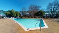 This community space combines a well-appointed indoor clubhouse with an inviting outdoor pool area. The pool is generously sized with crystal-clear turquoise water, surrounded by a textured concrete deck lined with lounge chairs. Mature trees frame the backdrop, creating a shaded, relaxed atmosphere under open skies.

Inside, the clubhouse is designed with an open-concept layout that flows seamlessly across rich hardwood flooring. Vaulted, coffered ceilings with crown molding lend architectural character and a sense of height throughout the space. Decorative white columns define distinct zones without closing off the layout.

A dedicated bar area features pendant lighting and patterned tile accents, complemented by bar-height seating along a built-in counter. A separate lounge zone includes a fireplace flanked by a mounted television and comfortable seating, ideal for relaxed gatherings. A high-top dining area with bistro-style stools provides additional flexible seating.

One section features a long wall-mounted counter with bar stools beneath a vaulted ceiling accent, functioning well as a co-working or event overflow space. The cool blue-gray walls throughout lend a calm, contemporary feel. Recessed lighting and large windows ensure the interior remains bright and welcoming.