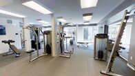 This fitness facility is designed to accommodate a comprehensive workout experience within a single-level layout. The space features a thoughtfully organized arrangement of exercise equipment, including cable resistance machines, a multi-function strength station, a weight bench, and cardio equipment from brands such as Precor. The flooring consists of durable, light-toned material suited for high-traffic use, with rubberized matting placed beneath key equipment zones for added safety and stability. Mirrored walls line one side of the room, enhancing the sense of depth and allowing users to monitor their form during exercise. The ceiling is finished in a clean white with recessed and flush-mounted lighting panels that ensure consistent, even illumination throughout the space. Large windows along the far wall invite natural light and offer views of the surrounding greenery, contributing to an open and energizing atmosphere. Structural columns are integrated into the layout without disrupting the flow of the room. Overall, the facility presents a functional, well-maintained environment suitable for a range of fitness routines.