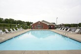This outdoor amenity space centers around a generously sized swimming pool with clear, light-colored water, providing a refreshing recreational focal point. The pool is complemented by a separate hot tub, offering additional relaxation options for residents. The surrounding concrete pool deck is spacious and well-maintained, lined with white lounge chairs and patio seating arranged beneath teal market umbrellas, creating comfortable shaded areas for lounging.

The space is enclosed by a sturdy black metal fence with gated entry points, providing security and controlled access. Adjacent to the pool deck stands a single-story brick building with classic red brick construction, white-trimmed multi-pane windows, and a pitched roof with decorative circular vents — lending the facility a traditional, well-kept architectural character. The exterior of this structure also features a mounted grill and waste receptacles for outdoor convenience.

Lush mature trees form a natural green backdrop beyond the fencing, enhancing the sense of privacy and creating a pleasant, park-like atmosphere. The surrounding grounds include open green lawn areas visible through the fence line. Overall, this amenity area is thoughtfully designed to offer a complete outdoor leisure experience in a clean, secure, and attractive setting.