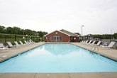 This outdoor amenity space centers around a generously sized swimming pool with clear, light-colored water, providing a refreshing recreational focal point. The pool is complemented by a separate hot tub, offering additional relaxation options for residents. The surrounding concrete pool deck is spacious and well-maintained, lined with white lounge chairs and patio seating arranged beneath teal market umbrellas, creating comfortable shaded areas for lounging.

The space is enclosed by a sturdy black metal fence with gated entry points, providing security and controlled access. Adjacent to the pool deck stands a single-story brick building with classic red brick construction, white-trimmed multi-pane windows, and a pitched roof with decorative circular vents — lending the facility a traditional, well-kept architectural character. The exterior of this structure also features a mounted grill and waste receptacles for outdoor convenience.

Lush mature trees form a natural green backdrop beyond the fencing, enhancing the sense of privacy and creating a pleasant, park-like atmosphere. The surrounding grounds include open green lawn areas visible through the fence line. Overall, this amenity area is thoughtfully designed to offer a complete outdoor leisure experience in a clean, secure, and attractive setting.