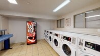 Located at 259 Colonnade Dr in Charlottesville, Virginia, this shared laundry facility is a single-floor space designed to serve the practical needs of its users efficiently. The layout is thoughtfully organized, with distinct zones for washing, drying, and waiting.

The washing area features a row of front-loading washing machines lined along one wall, providing ample capacity for multiple users simultaneously. Adjacent to the washers, a bank of stacked dryer units maximizes the use of vertical space, offering a generous number of drying options. Light tile flooring runs throughout the entire space, making for easy maintenance and a clean appearance.

Natural light filters in through several windows, including a larger window near the entrance that offers a view of the surrounding greenery. A glass-paned door provides access to the outdoors and contributes to the bright, open feel of the room. Overhead fluorescent lighting ensures the space remains well-lit at all times.

A dedicated waiting area features a row of chairs positioned near the entrance, offering a comfortable spot for users to relax while their laundry cycles run. A blue folding table provides a convenient surface for sorting and folding clothes. A Coca-Cola vending machine adds a convenient amenity for patrons. Hand sanitizer dispensers are also mounted on the walls throughout the space, promoting cleanliness and hygiene.