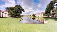 This residential community is characterized by its serene, park-like setting, defined by a central pond that serves as a focal point of the outdoor environment. The calm water reflects the sky and surrounding multi-story buildings, which feature warm earth-toned exteriors, red-tiled rooflines, and covered balconies — architectural details consistent with Florida-style residential design. The grounds are extensively landscaped with wide expanses of manicured green lawn, mature shade trees, and tropical plantings that add both privacy and visual appeal throughout the property.

The community's layout is spacious and open, with generous green areas between buildings that provide residents with room to enjoy the outdoors. The pond creates a naturally tranquil backdrop visible from multiple vantage points across the grounds. Palm trees and large canopy trees are distributed throughout, contributing to a lush, well-established atmosphere.

The unit itself is situated on a single floor within this larger community setting. While interior images were not sufficiently lit to capture room details, the overall environment suggests a well-maintained, nature-integrated residential setting with access to scenic outdoor views and ample communal green space.