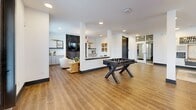 Located at 289 W Laughlin Ln in Celina, TX, this thoughtfully designed single-floor amenity space offers an open-concept layout that seamlessly connects a fully equipped kitchen, inviting lounge areas, and a dedicated game room. The kitchen is a standout centerpiece, featuring a long white island with brick-style paneling, bar seating, gray shaker cabinetry, a matte black faucet, stainless steel appliances, a beverage refrigerator, and a bold geometric patterned tile backsplash. Industrial-style pendant lights hang above the island, complementing the warm wood-look tile flooring that runs throughout the entire space. The living area exudes a relaxed yet refined ambiance, highlighted by a distinctive herringbone wood accent wall, a wall-mounted TV, a dramatic drum pendant chandelier, and curated artwork. Natural light pours in through multiple large windows. A separate game room area provides ample open floor space for a pool table and foosball table, framed by clean white walls adorned with eclectic artwork. Structural columns define zones within the open floor plan while maintaining a sense of spaciousness. A dining nook with a round table is tucked near the windows, completing this versatile and welcoming community retreat.