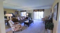 This single-floor living space offers a thoughtfully designed interior centered around a spacious, light-filled living area. A defining feature of the room is the large wall-to-wall mirror installation, which visually expands the space and amplifies natural light streaming in from the oversized sliding glass doors. These doors provide direct access to a private balcony, where outdoor seating can be glimpsed, extending the living area's functional footprint. Plush wall-to-wall carpeting runs throughout, contributing to a warm and comfortable atmosphere. The living area is generously proportioned, comfortably accommodating a full sofa ensemble, floral-patterned accent chairs, and a dark wood coffee table with decorative detailing. Elegant valance curtains in warm tones frame the windows, adding a refined touch to the overall aesthetic. A tall indoor plant near the balcony entrance introduces a natural element, complementing the bright, airy feel created by the abundant incoming daylight. The cohesive interior design, combining traditional furnishings with practical layout choices, creates a welcoming and functional living environment well-suited for everyday comfort.
