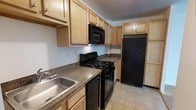 Unit 225-4S is a single-floor, 1-bedroom, 1-bathroom residence offering a functional and practical layout. The kitchen serves as a central feature of the unit, designed with efficiency in mind. It is outfitted with a full suite of black appliances, including a gas range, over-the-range microwave, dishwasher, and a full-size refrigerator. Light wood cabinetry lines both the upper and lower walls, providing generous storage space throughout. The countertops feature a dark granite-style laminate surface that contrasts neatly with the lighter cabinetry, while a stainless steel sink is conveniently positioned along the counter. The light-toned tile flooring adds brightness to the kitchen space. The galley-style layout of the kitchen maximizes the use of available space, keeping everything within easy reach. The overall design of the unit reflects a straightforward, practical aesthetic suited for comfortable everyday living, with well-maintained finishes throughout. The single-floor layout ensures easy accessibility and a cohesive flow between the living spaces.