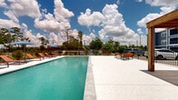 Located at 14441 T C Jester Blvd in Houston, Texas, this outdoor amenity space centers around a generously sized rectangular swimming pool with clear turquoise water. The pool deck is finished in a light-toned, textured surface that surrounds the pool on multiple sides, providing ample space for relaxation and movement. A row of reclining sun loungers lines one side of the pool, ideal for soaking up the Texas sun, while a shaded umbrella seating area offers a cooler retreat. On the opposite end, a wooden pergola provides additional shade and frames an outdoor seating arrangement with vibrant orange chairs and a central fire pit table, creating a comfortable lounge area. A built-in grill station is also present, making this space well-suited for outdoor gatherings. The pool area is enclosed by a wrought-iron fence, ensuring safety and privacy, with lush mature trees and open sky visible beyond the perimeter. Pool depth markers and posted safety signage are clearly visible throughout. The backdrop features a multi-story residential building in a bold navy blue exterior, reinforcing the community character of the property. The overall ambiance is open, bright, and inviting, designed for outdoor leisure and social activity.