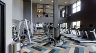 This fitness center in Dallas offers a thoughtfully designed workout space that combines functionality with contemporary style. The facility's most striking architectural feature is its soaring double-height ceiling, which creates an open, airy atmosphere and is complemented by large windows along the perimeter that invite abundant natural light throughout the day. Floor-to-ceiling mirrors are strategically positioned to visually expand the space while allowing users to monitor their form during exercise. The interior is anchored by a bold geometric-patterned carpet in teal, tan, and grey tones, adding a dynamic visual element underfoot. The equipment layout is well-organized, featuring a comprehensive selection of strength training machines and cable systems alongside a row of treadmills equipped with individual TV screens for entertainment during cardio sessions. Recessed and pendant lighting fixtures provide even illumination throughout the space. The overall color palette of charcoal grey walls paired with metallic equipment finishes gives the facility a sleek, modern aesthetic, making it a well-rounded and visually engaging environment for fitness enthusiasts.