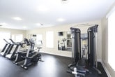 This dedicated fitness center offers a well-organized and fully equipped workout space designed to support a comprehensive exercise routine. The room features durable rubber flooring throughout, providing a safe and functional surface suited for a wide range of activities. Warm, neutral-toned walls paired with recessed lighting create a clean and energizing atmosphere conducive to focused training sessions.

The cardio area is outfitted with multiple treadmills and stationary bikes, including Cybex and BH branded machines, offering users a variety of aerobic options. A multi-functional cable machine and an adjustable weight bench round out the strength training offerings, allowing for a full-body workout without leaving the facility. Large wall-mounted mirrors run along one side of the room, visually expanding the space and allowing users to monitor their form during exercise.

Natural light filters in through multiple windows fitted with vertical blinds, adding brightness to the interior while maintaining privacy. A wall-mounted television is also present, offering entertainment during workouts. The layout is thoughtfully arranged to maximize usable floor space, ensuring comfortable movement between equipment. An adjacent room, visible through a glass-paneled door, suggests additional amenity space nearby, further enhancing the overall functionality of this fitness facility.