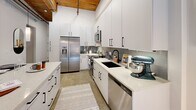This thoughtfully designed one-bedroom, one-bathroom loft-style unit showcases an impressive blend of industrial character and contemporary finishes across a single well-organized floor. The space is defined by striking architectural details, including exposed brick walls, polished concrete floors throughout, and warm wood-beam ceilings that lend a distinctive urban aesthetic.

The kitchen is a standout feature, offering an expansive galley-style layout with floor-to-ceiling white cabinetry accented by matte black hardware, a gray tile backsplash, white quartz countertops, and a full suite of stainless steel appliances including a French door refrigerator, gas range, microwave, and dishwasher. A kitchen island provides additional workspace and storage.

The bedroom is an intimate retreat framed by a large arched window that fills the space with natural light and outdoor views. Exposed brick and a raw concrete column add to the room's industrial character. The living room offers a cozy nook with carpeted flooring, providing a softer contrast to the concrete found elsewhere.

The bathroom is clean and well-appointed, featuring white subway tile surrounding a full soaking tub and shower, a white vanity with black hardware, and polished concrete flooring. The layout also includes a dedicated entrance, hallway, and closet, ensuring practical everyday functionality throughout.