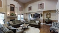 This single-floor space makes an immediate impression with its dramatic, soaring ceilings that lend an open and airy atmosphere throughout. The main lounge area is thoughtfully appointed with plush, tufted sofas arranged around a round marble-top coffee table, all anchored by a large oval area rug over warm hardwood flooring. Decorative wall panels and carefully placed artwork add visual interest at multiple heights, complementing the neutral, beige-toned color palette that gives the room a calm and inviting character.

A classic fireplace anchors one end of the lounge, framed by traditional millwork and flanked by tall French doors that invite natural light deep into the space. On the opposite side, a built-in coffee bar is neatly recessed into the wall beneath an arched surround, complete with cabinetry and a coffee machine — a convenient amenity for guests and visitors. Wall-mounted floral arrangements and sconce lighting add decorative warmth throughout.

The overall ambiance is refined yet comfortable, blending traditional architectural details — such as crown molding and columned archways — with practical, welcoming furnishings. The space is well-suited for relaxed socializing or quiet rest, with abundant seating and a cohesive, elegantly understated design sensibility.