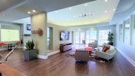 "The Hub" is a thoughtfully designed communal lounge space that blends contemporary style with a welcoming atmosphere. The single-floor layout features rich hardwood flooring throughout, complemented by soft sage-green walls and crisp white trim that create a clean, modern aesthetic. Recessed lighting is complemented by a striking Sputnik-style chandelier, lending a sophisticated focal point to the main seating area. A large flat-screen television is mounted above a sleek media console, making the space well-suited for both casual viewing and social gatherings. The seating arrangement includes plush gray lounge chairs, a bold red accent ottoman, and a patterned area rug that anchors the space with visual interest. Generous floor-to-ceiling windows and glass-paneled double doors flood the interior with natural light while offering views of the surrounding greenery outdoors. Architectural detailing includes a tray ceiling that adds depth and dimension to the room. A tall potted plant near the entrance introduces a natural element, softening the modern design. Overall, The Hub presents a polished, inviting environment ideal for community gathering and relaxation.