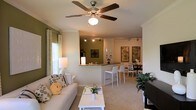 This single-floor residence showcases an open-concept floor plan that seamlessly connects the living room, kitchen, and dining area into one cohesive, functional space. The living room is comfortably scaled, featuring plush seating, a white coffee table, and neutral carpet flooring that runs throughout. Crown molding lines the ceilings, and a ceiling fan with a built-in light fixture serves as a central focal point overhead. Natural light enters through a window dressed with patterned curtains in green and white, complementing the warm olive-green accent wall that anchors the kitchen area. The kitchen features a raised breakfast bar with white bar stools, offering a casual seating option and a natural visual divider between the cooking and living spaces. Track lighting above the kitchen provides focused illumination. The dining area is situated just beyond, accommodating a dark wood table and chairs beneath framed botanical artwork. The overall ambiance is warm and inviting, with a cohesive color palette of greens, beiges, and whites tying each zone together.
