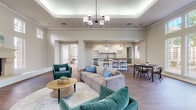 This elegant single-floor communal space is designed to impress, combining classic architectural details with a warm, inviting atmosphere. The main lounge area features soaring ceilings with a coffered tray detail, accented by an eye-catching modern chandelier that serves as a focal point overhead. Rich hardwood-style flooring runs throughout, anchoring the open, flowing layout with a sense of continuity and warmth.

A striking floor-to-ceiling fireplace with a refined stone surround anchors one end of the lounge, flanked by tall windows that bathe the space in abundant natural light and offer views of the surrounding greenery. Teal velvet armchairs and a neutral sectional sofa are arranged around a round wooden coffee table, creating a welcoming conversation area.

The space transitions naturally into a dining zone, where multiple seating arrangements accommodate groups comfortably. Large multi-pane windows along one wall flood the dining area with daylight and provide a pleasant outdoor vista. Adjacent to the dining area, a sleek kitchen bar with white cabinetry, pendant lighting, open shelving, and counter-height seating offers a casual yet functional gathering spot. A separate room visible from the lounge appears to serve as a private workspace or office area, adding to the space's versatility.