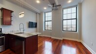 This single-floor urban residence offers a thoughtfully designed open layout highlighted by rich cherry hardwood flooring that flows throughout the main living areas. The open-concept living and kitchen space is anchored by floor-to-ceiling industrial-style windows that flood the interior with natural light while framing sweeping city views. A ceiling fan complements the airy atmosphere created by the elevated ceiling height, finished with classic crown molding.

The kitchen is a standout feature, showcasing deep mahogany cabinetry, a large center island clad in a rich panel finish, and elegant marble-veined countertops. Stainless steel appliances, including a refrigerator, built-in microwave, and gas range, are paired with a sleek stainless hood and a modern pendant light fixture, making the space as functional as it is visually striking.

A dedicated in-unit laundry closet houses a stacked washer and dryer, adding everyday convenience. The bedroom offers neutral carpet, crown molding, and a spacious reach-in closet. The bathroom is finished with large-format gray marble-style tile, a glass-enclosed shower, a dark-framed vanity with a white countertop, and polished chrome fixtures throughout.