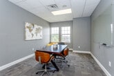 Located at 1401 College Ave in Manhattan, Kansas, this single-floor professional space offers a well-appointed meeting room designed for productivity and collaboration. The room is finished with neutral gray walls and patterned carpet tiles, creating a clean and focused atmosphere. A dark wood conference table serves as the centerpiece, surrounded by stylish mid-century-inspired rolling chairs with bentwood backs, providing comfortable seating for small group meetings or discussions. Natural light filters in through multiple white-framed windows along one wall, offering views of the surrounding outdoor environment. A large glass whiteboard is mounted on one wall, making the space well-suited for brainstorming sessions and presentations. A decorative world map artwork adds a global, professional touch to the room's aesthetic. A standout architectural feature is the interior window that offers a direct sightline into an adjacent game room, visible with a pool table, adding a unique and inviting character to the overall space. The suspended acoustic tile ceiling with recessed lighting ensures even illumination throughout. This space balances a professional, functional design with subtle creative elements, making it suitable for a variety of collaborative or business-oriented uses.