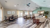 Located at 8799 Old Colony Way in Alexandria, VA, this single-floor space offers a thoughtfully designed layout that combines comfort and functionality. The living room is the centerpiece of the space, characterized by dramatic vaulted ceilings with dual ceiling fans and recessed lighting that create an airy, open atmosphere. Rich, multi-toned hardwood-style flooring runs throughout, anchoring the warm neutral color palette of the walls and furnishings. A white-mantled fireplace with a tile surround serves as a striking focal point, flanked by windows that allow abundant natural light to fill the room. Multiple seating arrangements provide versatile areas for relaxing and entertaining, while a dedicated dining area adds to the functionality of the living space. An accent wall in soft teal adds a subtle pop of color. The kitchen features warm, honey-toned wood cabinetry paired with granite-look countertops, white appliances including a range with over-the-range microwave, and ample storage throughout. Large windows and glass doors throughout the space frame outdoor views, connecting the interior to the surrounding environment and enhancing the overall sense of openness and light.