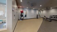 Located at 925 Clopper Rd in Gaithersburg, Maryland, this single-floor fitness center offers a comprehensive workout facility designed to support a wide range of exercise routines. The space is thoughtfully laid out across several interconnected zones, each serving a distinct fitness purpose.

The main cardio and strength area features an extensive lineup of equipment, including treadmills, elliptical trainers, stationary bikes, and a variety of Hoist-brand selectorized strength machines. Large windows along the perimeter flood the space with natural light, creating an energizing atmosphere. Ceiling fans and recessed lighting complement the bright, open feel throughout.

A dedicated free weights section houses a dumbbell rack alongside open floor space, supported by recessed lighting and a clean, neutral color palette. Adjacent to this area is a water fountain and a built-in wooden shelving unit, providing convenient storage for personal belongings.

A separate functional training and stretching studio offers open floor space finished with rubberized flooring in a two-tone design. This room is appointed with stability balls, medicine balls, and motivational fitness-themed wall art and typography, reinforcing an encouraging workout environment.

The flooring throughout the facility consists of durable rubber gym flooring, suitable for both cardio and strength training. Glass partition walls divide sections while maintaining visual openness. The overall design is modern, clean, and purpose-built for an effective fitness experience.