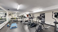 Located at 2001 Carling Ave in Ottawa, Ontario, this fitness facility offers a practical and well-equipped workout space across a single floor. The gym is organized to maximize functionality, with the main exercise area housing a lineup of cardio machines — including multiple treadmills, an elliptical trainer, a stationary bike, and a stair climber — arranged to allow ease of movement between equipment. A large wall mirror on one side visually expands the space and supports proper form during exercise. Free weights, resistance bands, and exercise mats are also available, catering to a range of workout preferences. Motivational black-and-white fitness photography adorns the walls, contributing to an energizing atmosphere. The flooring throughout is durable rubber, suitable for high-traffic fitness use. Overhead fluorescent lighting provides consistent, bright illumination across the entire space. A secondary area at one end of the facility features a wall-mounted flat-screen television, a water fountain with a bottle-filling station, a paper towel dispenser, and waste receptacles — offering convenient amenities for users during and after their workout. Baseboard heating ensures year-round comfort. The facility is accessed via a secured door, providing a private and focused environment for exercise.
