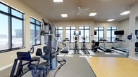 This fitness center is designed to support a well-rounded workout experience. The space is outfitted with a multi-station strength training machine positioned prominently near the entrance, offering a range of resistance exercise options. Beyond it, a dedicated cardio zone features several treadmills, an upright stationary bike, and a rowing machine, providing diverse aerobic training options. Mounted televisions are positioned for easy viewing during workouts. The flooring combines neutral carpet in the exercise areas with a hardwood section, delineating functional zones within the room. Large windows line multiple walls, flooding the space with natural light and offering open views of the surrounding exterior. A ceiling fan overhead aids in air circulation, contributing to a comfortable workout environment. The walls are finished in a neutral beige tone, and recessed lighting supplements the natural light. The overall atmosphere is practical and inviting, with a clean, organized layout that accommodates both strength and cardiovascular training needs efficiently.