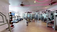 Located at 13330 Noel Rd in Dallas, TX, this fitness center is a single-floor exercise facility offering a thoughtfully designed workout environment. The space features light wood-look flooring throughout, white walls, and a distinctive pink LED neon lighting installation that traces a curved path across the ceiling, adding a modern, energetic ambiance to the room.

The main workout area is generously proportioned and open, housing an extensive array of equipment. Cardio machines — including treadmills and stair climbers — are positioned along one side near windows that allow natural light to filter in. A multi-station cable and strength training machine serves as a central fixture, complemented by a full dumbbell rack and additional resistance machines along the mirrored walls. The mirrors visually expand the space while allowing users to monitor their form.

A dedicated area features a spin bike with an integrated digital display, stability balls on a vertical rack, and a large flat-screen TV mounted against a textured, geometric-patterned accent wall — ideal for guided virtual workouts. A medicine ball rack and a chest press machine are positioned near the entrance area, which also includes a hand sanitizer station and posted fitness rules. Ceiling fans provide additional ventilation, making this a fully functional and well-appointed fitness facility.