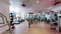 Located at 13330 Noel Rd in Dallas, TX, this fitness center is a single-floor exercise facility offering a thoughtfully designed workout environment. The space features light wood-look flooring throughout, white walls, and a distinctive pink LED neon lighting installation that traces a curved path across the ceiling, adding a modern, energetic ambiance to the room.

The main workout area is generously proportioned and open, housing an extensive array of equipment. Cardio machines — including treadmills and stair climbers — are positioned along one side near windows that allow natural light to filter in. A multi-station cable and strength training machine serves as a central fixture, complemented by a full dumbbell rack and additional resistance machines along the mirrored walls. The mirrors visually expand the space while allowing users to monitor their form.

A dedicated area features a spin bike with an integrated digital display, stability balls on a vertical rack, and a large flat-screen TV mounted against a textured, geometric-patterned accent wall — ideal for guided virtual workouts. A medicine ball rack and a chest press machine are positioned near the entrance area, which also includes a hand sanitizer station and posted fitness rules. Ceiling fans provide additional ventilation, making this a fully functional and well-appointed fitness facility.