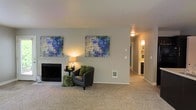 This single-floor residence offers a thoughtfully arranged living space designed for comfort and functionality. The main living area is a standout feature, showcasing a classic tiled fireplace that serves as a natural focal point of the room. Soft, wall-to-wall carpeting in a warm neutral tone runs throughout, creating a cozy and inviting atmosphere underfoot. The walls are painted in a cool, muted gray that complements the overall aesthetic and provides a versatile backdrop for décor. Warm ambient lighting from a stylish table lamp and a ceiling fixture adds a welcoming glow to the space. Two vibrant abstract art pieces in shades of blue, purple, and white add a creative touch to the walls, infusing the room with color and personality. A hallway visible from the living area leads to additional rooms, suggesting a well-connected and accessible floor plan. The corridor is illuminated with a ceiling-mounted light, hinting at further living spaces beyond. The overall ambiance of this residence is calm and contemporary, with a balanced mix of neutral tones and warm lighting that makes the space feel both functional and comfortable.