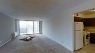This single-floor unit offers a clean, functional layout designed to maximize open living space. The main living area is generously sized, finished with wall-to-wall neutral carpeting that adds warmth underfoot. A striking wall of floor-to-ceiling sliding glass doors anchors one end of the room, flooding the interior with natural light and providing direct access to a private balcony with views of the surrounding urban landscape and neighboring buildings. The expansive white walls create a bright, airy atmosphere, offering a versatile blank canvas for a variety of interior styles. A textured ceiling adds subtle visual character to the space. Adjacent to the main living area, a clearly defined corridor leads to additional rooms, suggesting a practical and well-organized floor plan. The overall ambiance is light, open, and understated, with an emphasis on clean lines and uncluttered spaces. The combination of abundant natural light, open layout, and balcony access makes this unit a comfortable and adaptable living environment suitable for a range of uses and furnishing preferences.