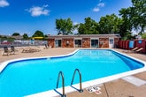 This single-story shared amenity space provides residents with convenient, well-maintained communal facilities. The centerpiece of the outdoor area is a generously sized rectangular swimming pool with clear blue water, enclosed by a red wooden privacy fence. Lounge chairs are positioned along the pool deck, and mature trees provide natural shade in the background, creating a relaxed outdoor retreat during warmer months.

The interior laundry facility is thoughtfully laid out and spacious, featuring a row of multiple top-loading washing machines aligned along one wall, paired with corresponding dryers, including stacked units near the entrance. The room is finished with light blue walls, accented by a decorative floral-patterned wall display that adds a welcoming touch. Tile flooring, overhead fluorescent lighting, and a window allowing natural light contribute to a clean, functional environment. A seating area with a small table and chairs offers a convenient waiting space.

Adjacent to the laundry room, a common entry area includes a bank of individual resident mailboxes, providing a centralized mail retrieval point. Safety features such as a fire extinguisher are visibly present. Together, these amenities reflect a practical, community-oriented facility designed to support daily residential needs.