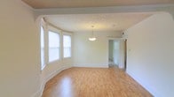 This single-floor space offers a clean, open layout filled with natural light and thoughtful architectural details. The main living area showcases warm hardwood flooring that runs seamlessly throughout, complemented by crisp white walls and classic crown molding that add a timeless, refined character to the space. A charming bay window serves as a focal point, flooding the room with generous natural light while offering an inviting sense of openness. Overhead, a simple yet elegant pendant light fixture adds a decorative touch to the living area. The textured ceiling adds subtle dimension, contributing to the room's overall warmth and character. Wide baseboards frame the space neatly, reinforcing the clean lines and traditional styling found throughout. An open doorway leads to an adjacent room, suggesting a functional and connected floor plan that allows for easy flow between spaces. The neutral color palette and well-maintained finishes create a bright, move-in-ready environment that provides an excellent canvas for a variety of interior design styles. Overall, this space balances classic architectural charm with practical, light-filled living areas.