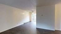 This single-floor space is defined by a clean, open layout that emphasizes functionality and simplicity. The main living area is generously proportioned, featuring dark hardwood-style flooring that contrasts sharply with the bright, white walls to create a crisp and inviting atmosphere. A textured popcorn ceiling adds a classic touch overhead, while a ceiling fan with an integrated light fixture provides both air circulation and ambient illumination.

Natural light flows into the space through a large window at the far end of the living area, offering a view of an outdoor fenced area and enhancing the sense of openness. Baseboard heating runs along the lower portion of the walls, ensuring efficient and consistent warmth throughout the space.

The walls are painted in a neutral off-white tone, providing a versatile backdrop that complements a wide range of interior design styles. The open floor plan allows for flexible furniture arrangement, making the living area highly adaptable. Overall, the space presents a straightforward, well-lit, and practical environment suited for comfortable everyday living.